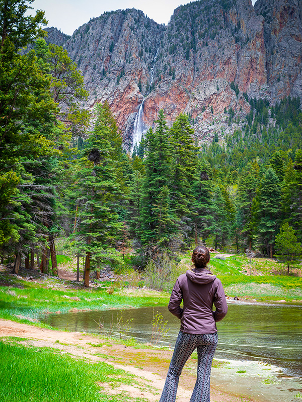 Woman looking at pond and pine trees with mountain cliffs and waterfall in background