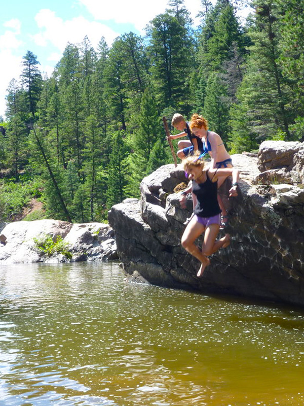 Woman and children jumping off cliff into river swimming pool with pine trees and boulders in background