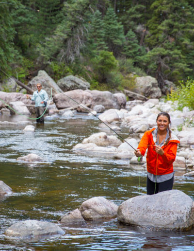 Woman in orange jacket and man fly fishing in waders in river with boulders and pine trees