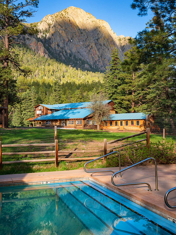 Outdoor swimming pool in foreground with wood cabin in background with mountain and pine trees