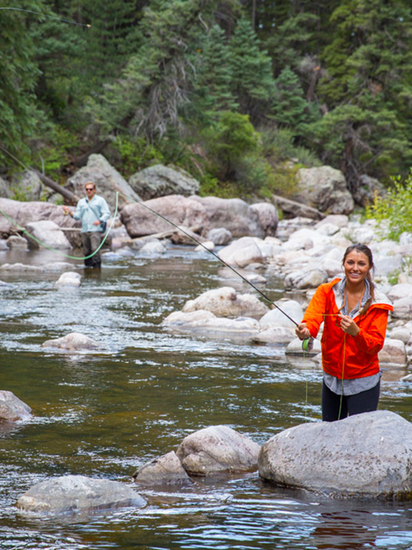Woman and man fly fishing in the river with boulders and pine tree forest in background
