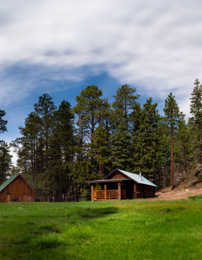 Outside view of grassy meadow and log cabins with pine trees in background and blue cloudy sky