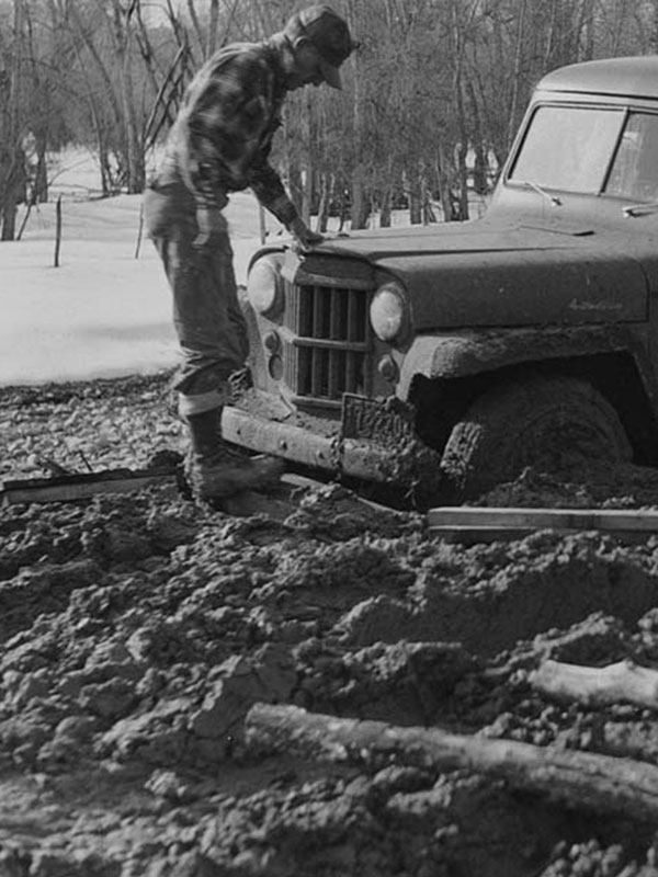 Black and white photo of old truck from the 40s in the mud in winter with man in plan shirt looking at it
