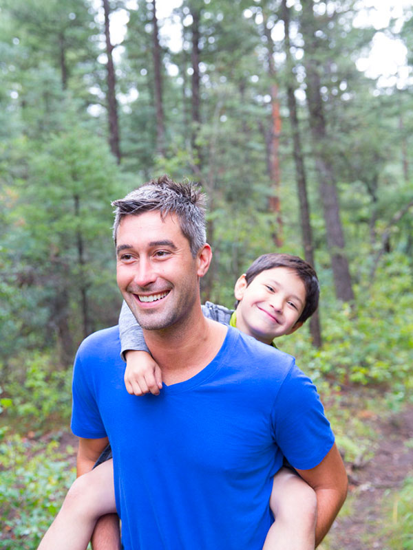 Dad in blue t-shirt with son on piggy back hike through forest trees