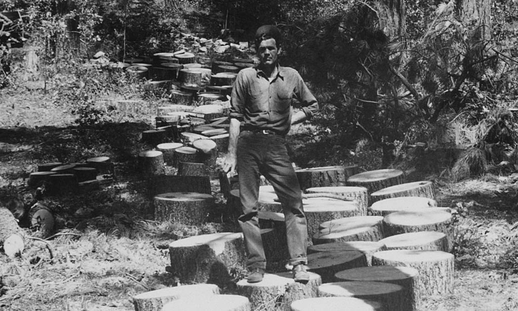 Black and white photo from the 1920s of white man standing on logging rounds in the mountains