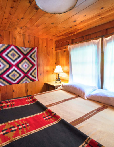 Bedroom with bed and red and blue bedspread wooden walls and ceiling with decorative rug on walls, large windows over bed