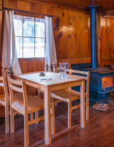 Wooden kitchen table and chairs next to wood burning stove in wood lined cabin