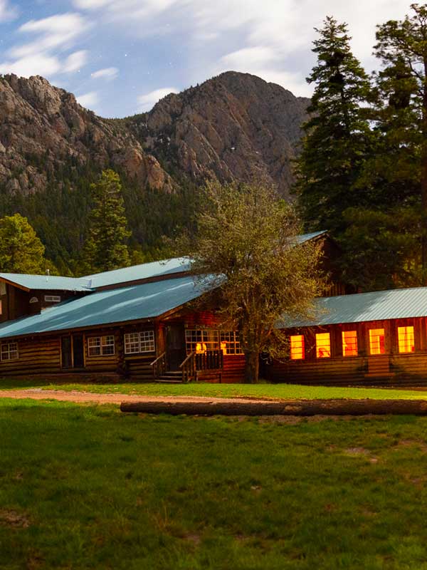 Old Lodge and game room building with lit interiors, grassy meadow and mountain and pine trees in background
