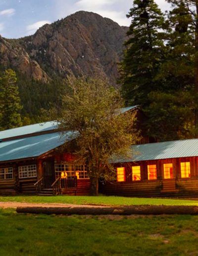 Log cabin building with lights in windows, grassy meadow and mountains in background