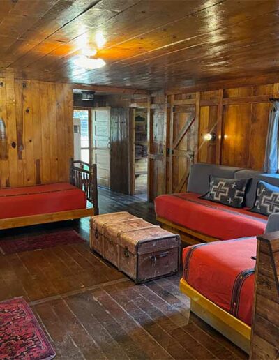 4 Twin beds in a living room area with a trunk in the middle and wooden ceiling, walls and floor.
